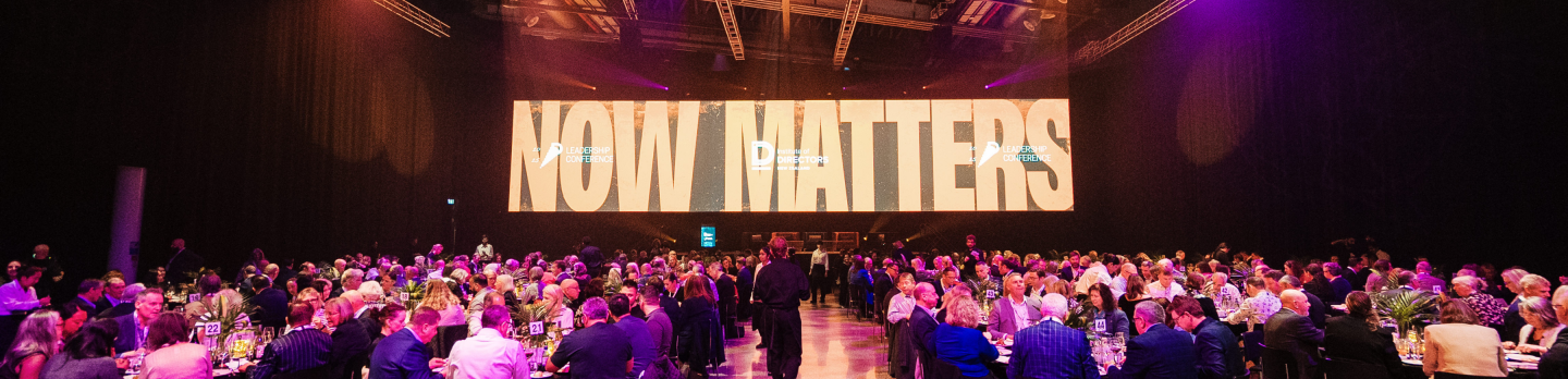 A large dining event with numerous guests seated at round tables under purple lighting. A screen displays the words "NOW MATTERS" in bold.
