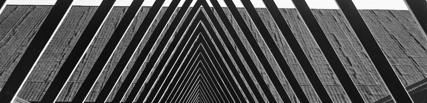 Black and white image of a series of converging triangular metal beams forming a geometric tunnel, creating a sense of depth and perspective against a textured wall.