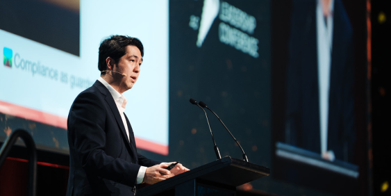 A suited man speaks at a conference, addressing attendees with confidence and clarity.
