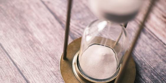 An hourglass filled with sand sits on a wooden table, casting a soft shadow in the ambient light.
