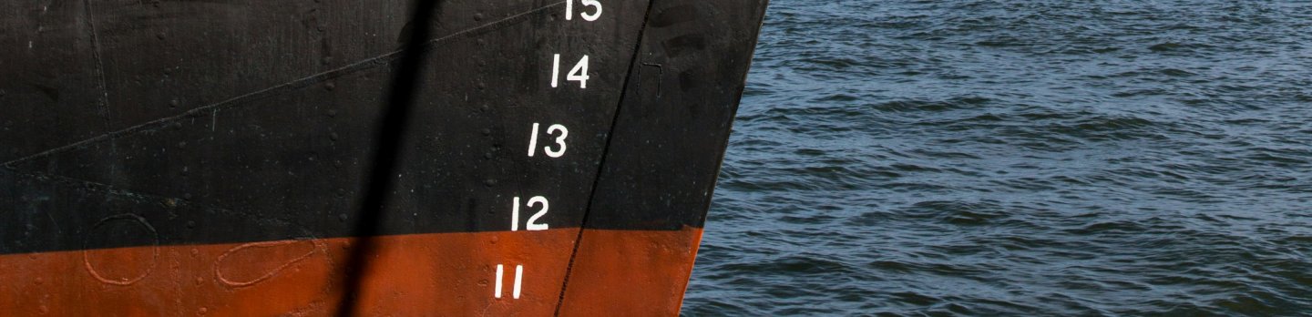 The image shows the bow of a ship with white draft numbers from 10 to 15 against a black and red-hulled background, adjacent to rippling blue water.