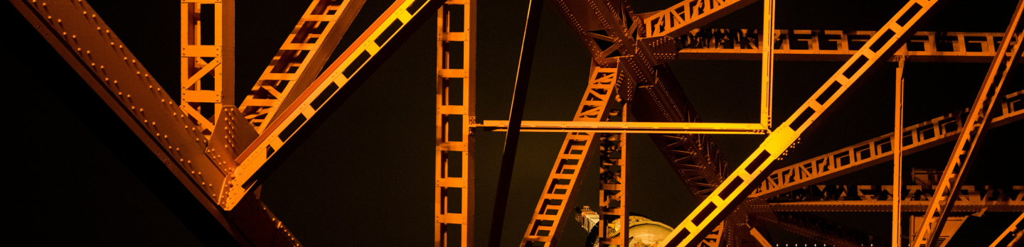 Intricate steel framework of a bridge, illuminated in orange light against a dark sky. The geometric patterns create a striking, industrial aesthetic.