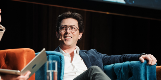 A man with glasses sits comfortably on a blue chair, appearing engaged in thought.