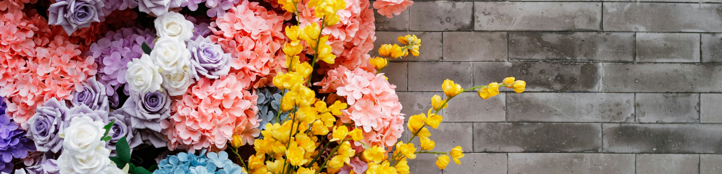 A vibrant floral wall with clusters of pink, purple, blue, and yellow flowers against a gray brick background, creating an uplifting, colorful display.