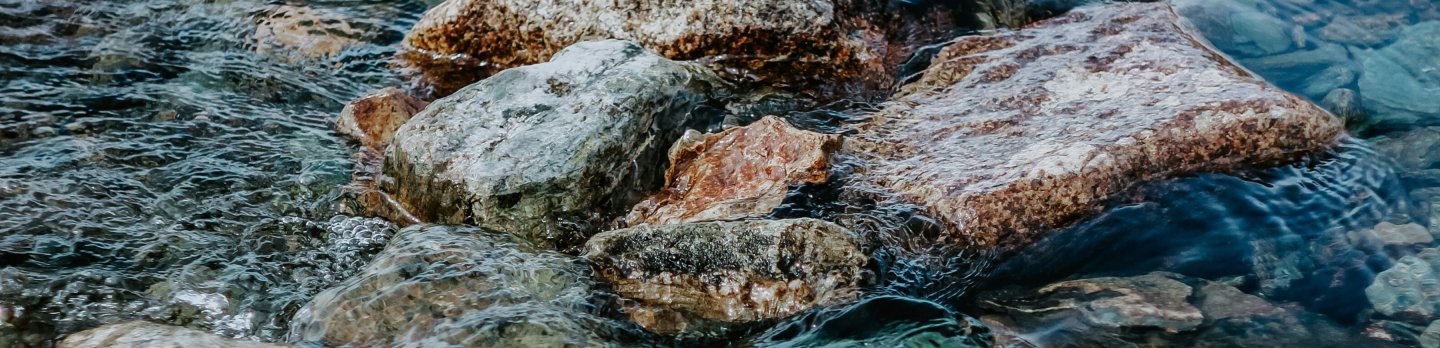 Clear mountain stream flows over smooth, multicolored rocks, with rippling blue-green water and gentle movement creating a calm scene.