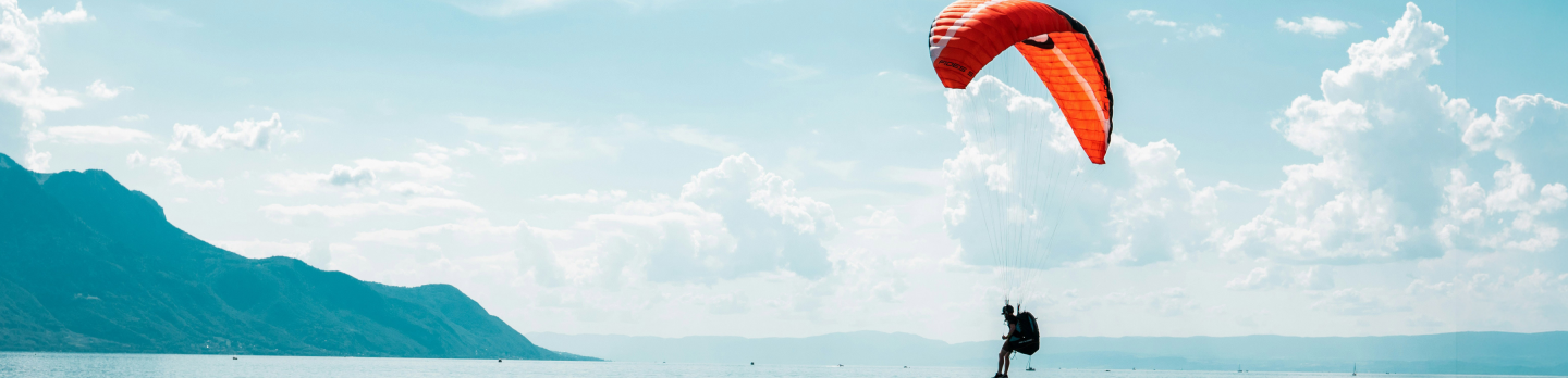 A paraglider with an orange chute soars over calm water near a floating platform.