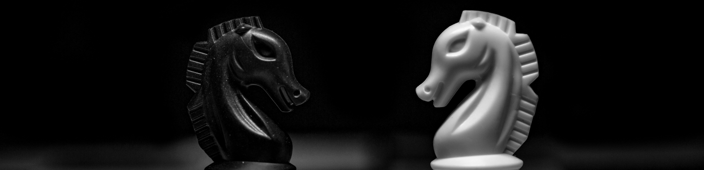 Black and white chess knights face each other on a dark board, their sculpted horse heads lit against a black background, creating a dramatic, tense mood.