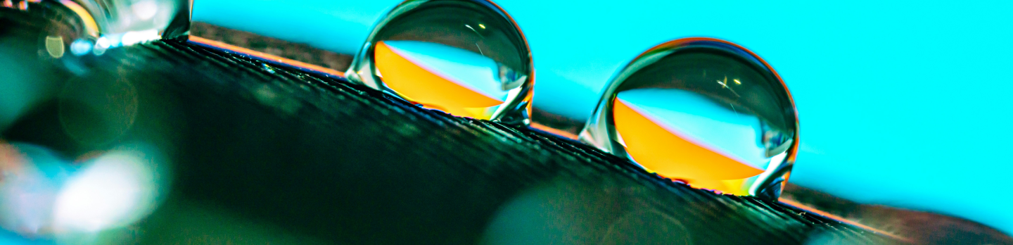 Macro photo of three clear water droplets balanced on a dark, curved leaf, reflecting bright yellow and orange light against a vivid turquoise background.
