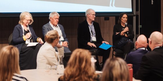 Four panelists sit onstage at a conference, with a woman speaking into a microphone as the audience listens; the mood is thoughtful and professional.