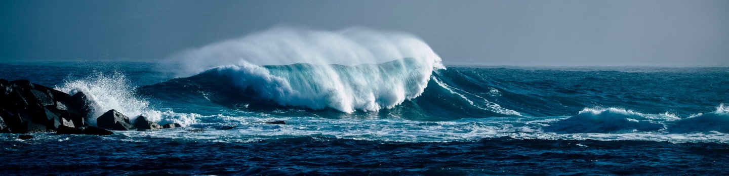 A large wave crashes on the ocean, with a majestic mountain rising in the background under a clear blue sky.