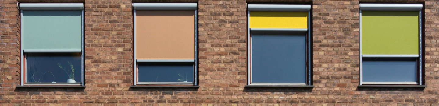 Four windows in a brick wall show varied roller blinds—two blue-gray, one tan, and one green—partly lowered, creating a simple, orderly architectural scene.
