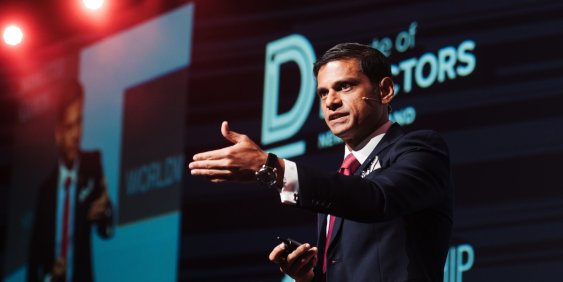 A confident male speaker in a dark suit gestures toward the audience on a blue-lit conference stage, beneath red spotlights and a technology-themed backdrop.
