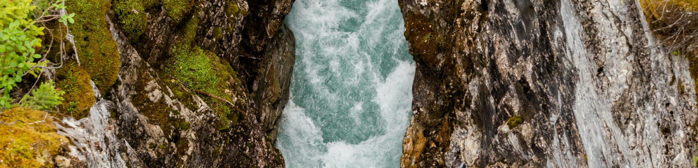 Turquoise river rapids rush through a narrow rocky gorge, bordered by moss-covered cliffs and green trees, creating a dramatic, untamed wilderness scene.