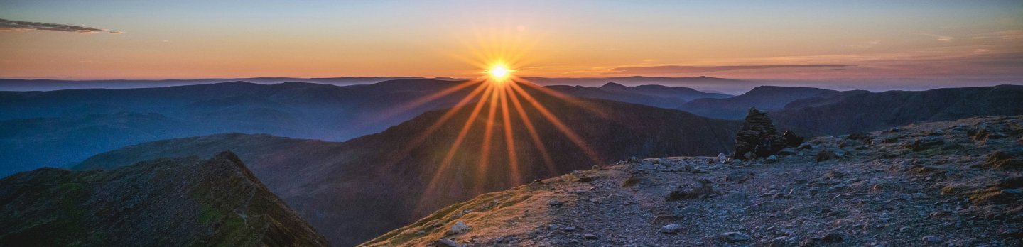 Sunrise breaks over layered mountain ridges, casting golden rays across a rugged rocky summit beneath a clear blue sky; the scene feels calm and awe-inspiring.