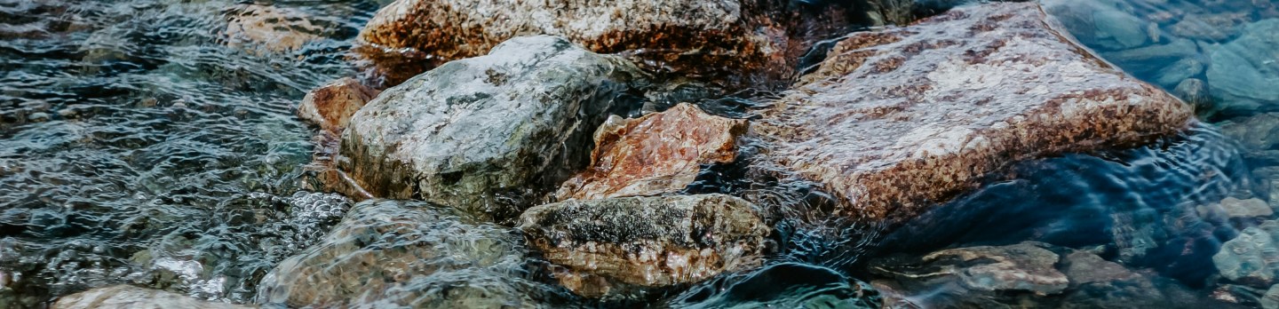 Clear mountain stream flows over smooth, multicolored rocks, with rippling blue-green water and gentle movement creating a calm scene.