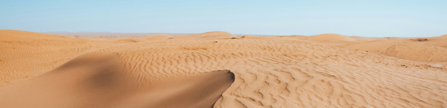 A sweeping desert dune with dark, smooth sand beside pale, rippled ridges beneath a vast clear blue sky, conveying quiet and isolation.