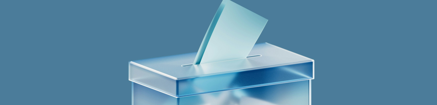 A transparent ballot box with papers inside sits on a pedestal against a blue background. One blue paper is partially inserted, indicating voting.
