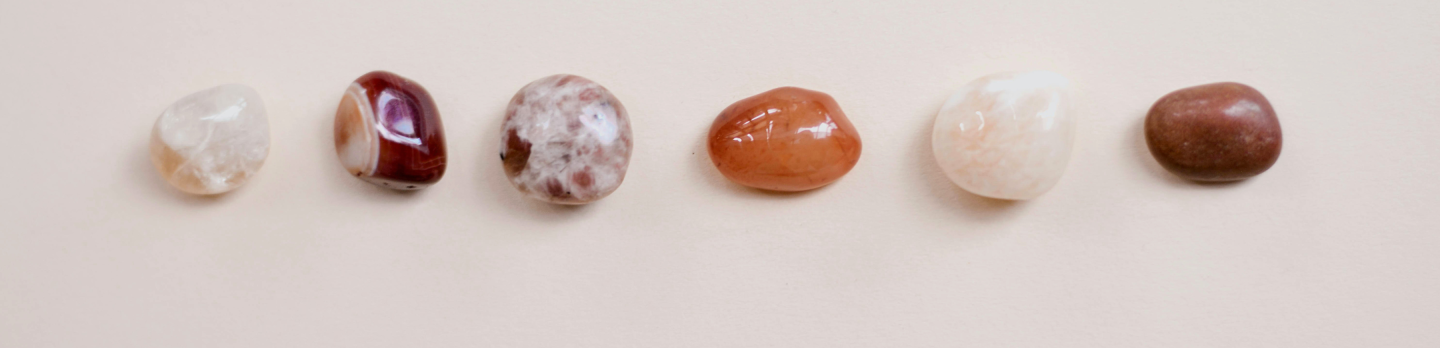 Six polished stones in varied colors, including white, red, and brown, are evenly spaced on a smooth, neutral beige background. 
