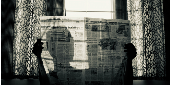 A person stands by a window, holding a newspaper in front of them, with natural light illuminating the scene.