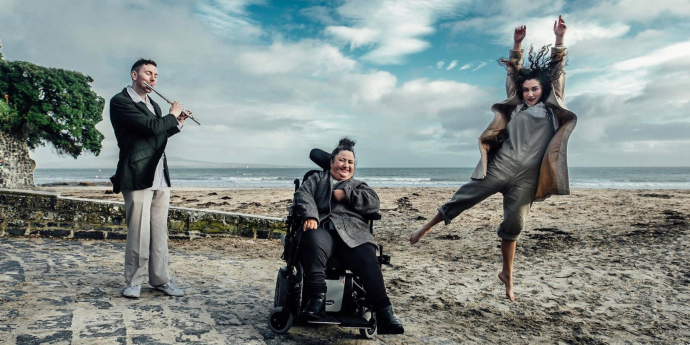 Three people on the beach, one playing the flute, one dancing and the other sitting in a wheelchair