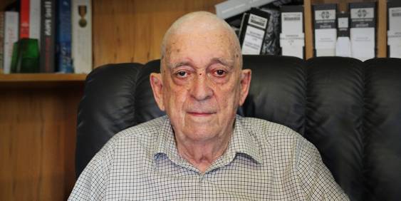Hugo Bedfort - White male in his 80's sitting on a black chair in front of a bookcase